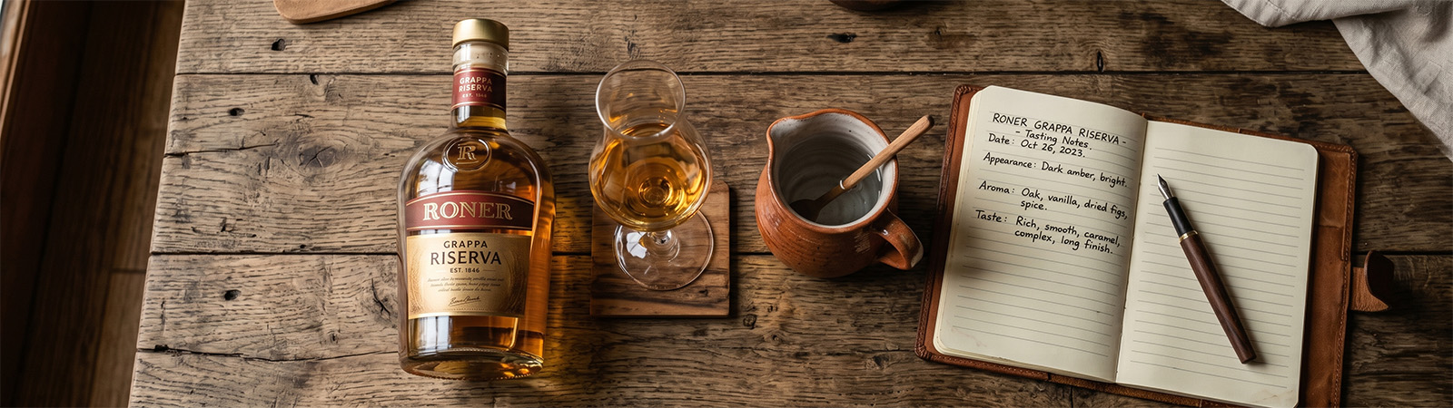 Vogelperspektive auf einen rustikalen Holztisch mit einer Flasche Roner Grappa Riserva, einem gef�llten Nosing-Glas auf einem h�lzernen Untersetzer, einem Tonkrug mit einem kleinen L�ffel und einem offenen ledernen Notizbuch mit handgeschriebenen Verkostungsnotizen und einem F�llfederhalter.