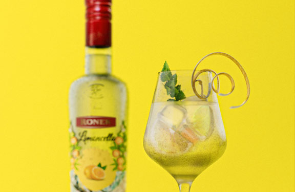 Roner Limoncello Flasche auf gelbem Hintergrund mit Glas und Deko im Vordergrund